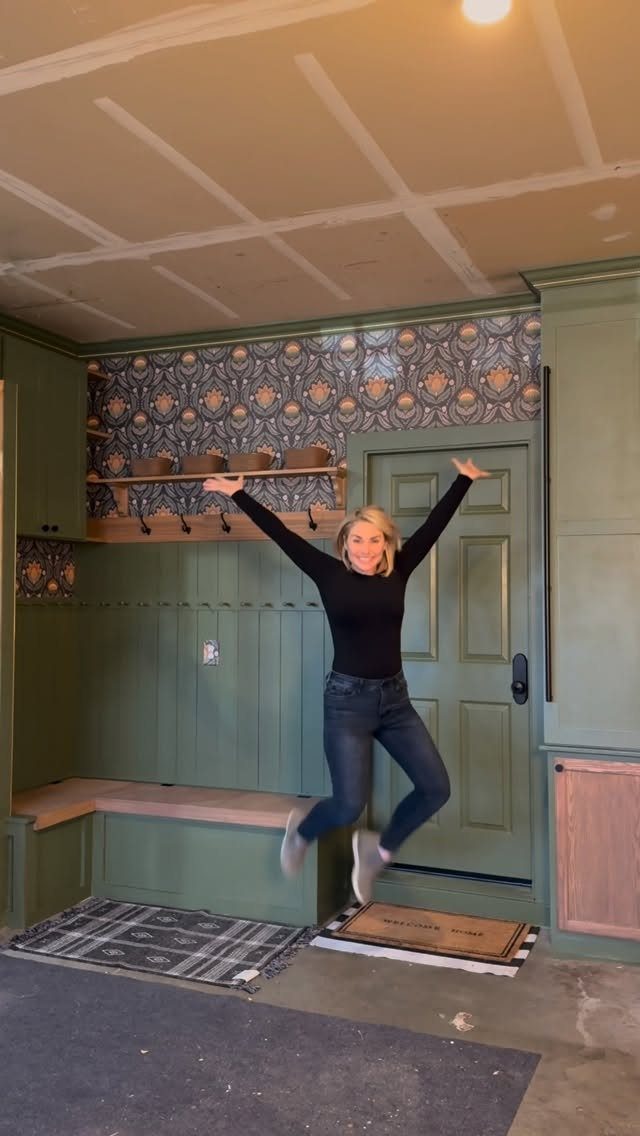 2 Car Garage Makeover Ideas: 8 Projects for a Finished Space 6 Woman jumping in front of a custom green mudroom with built-in bench and patterned wallpaper.