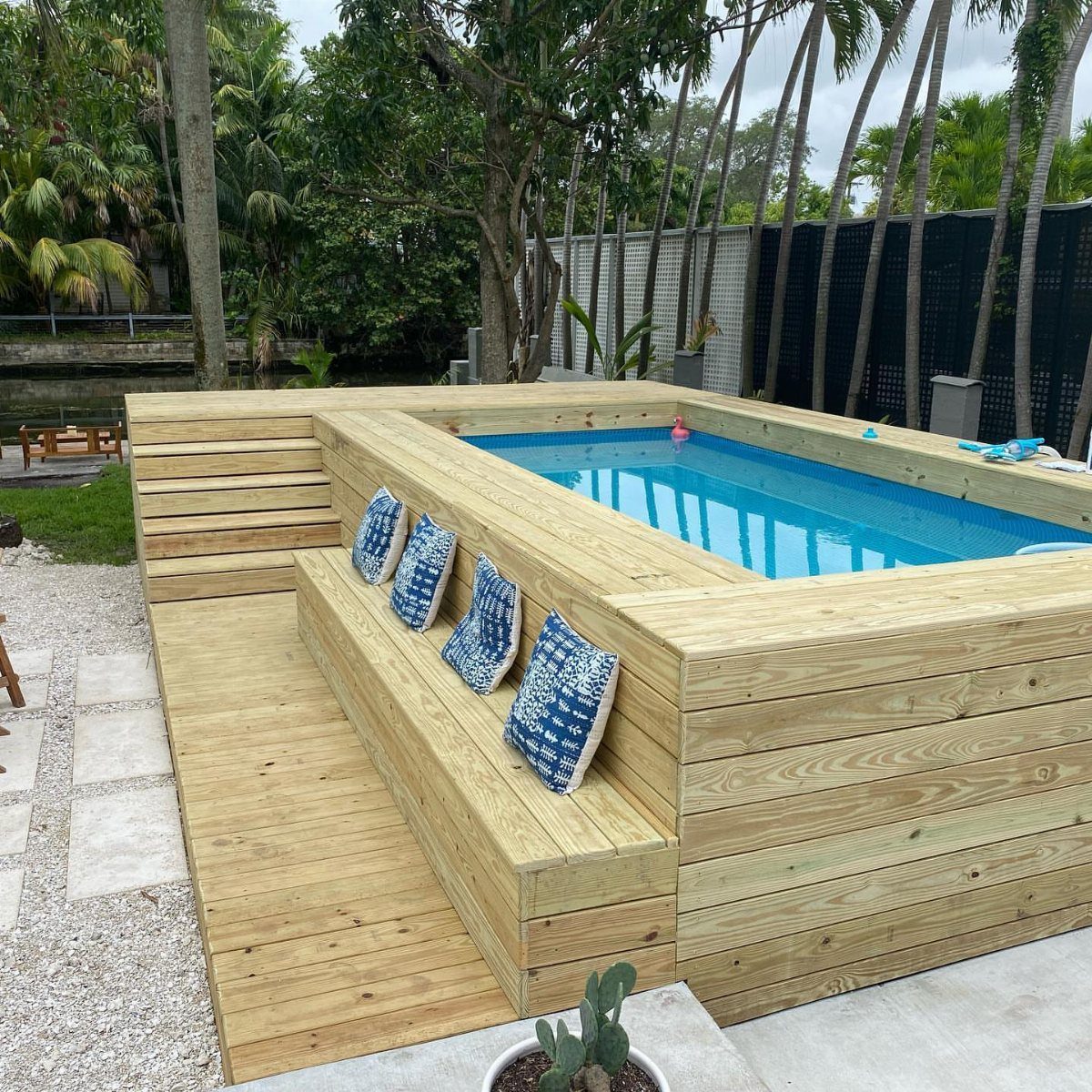 Wooden deck with built-in benches and blue pillows surrounding a rectangular swimming pool.