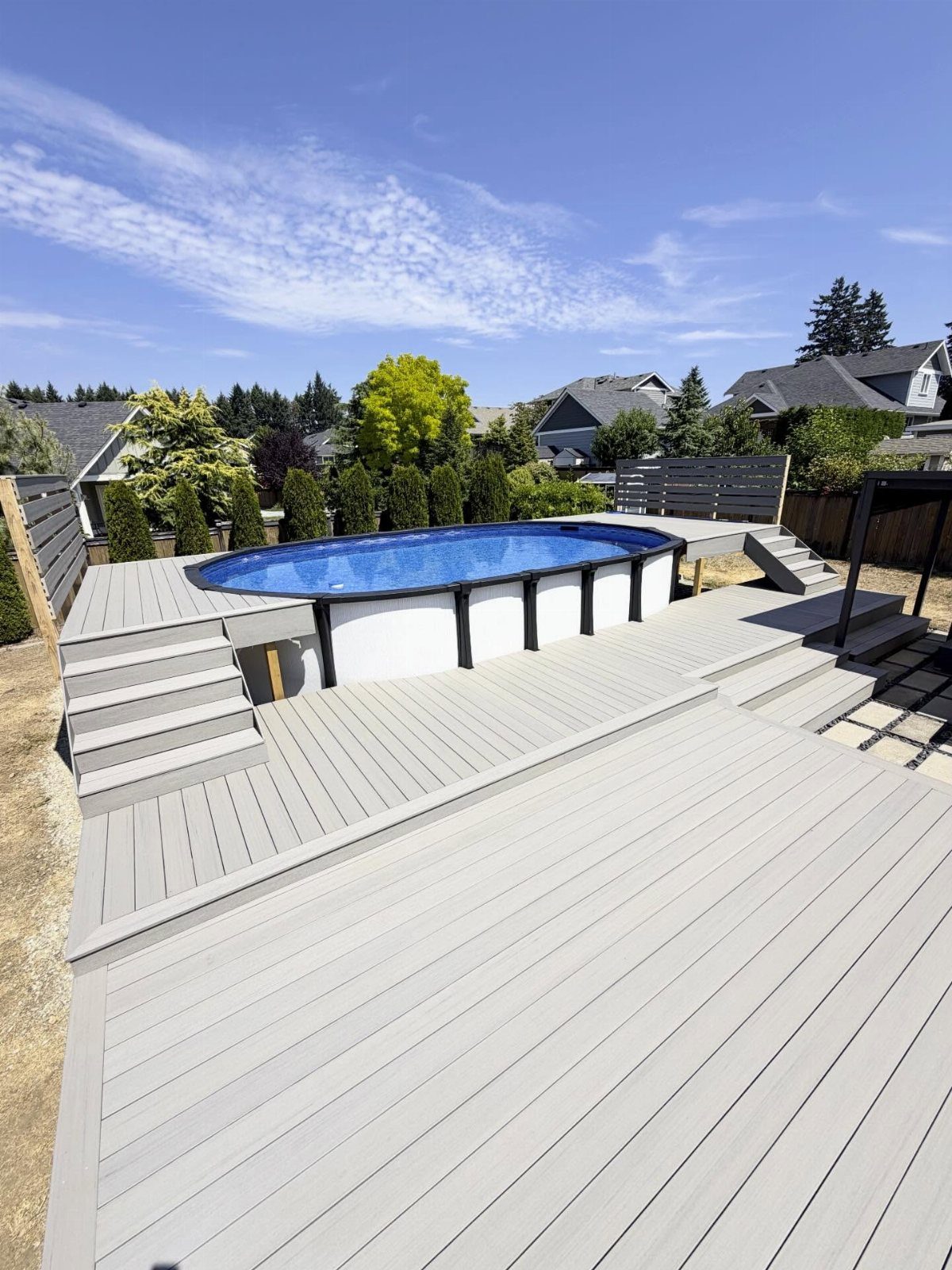Grey multi-level composite deck surrounding an oval blue above-ground pool in a backyard setting.