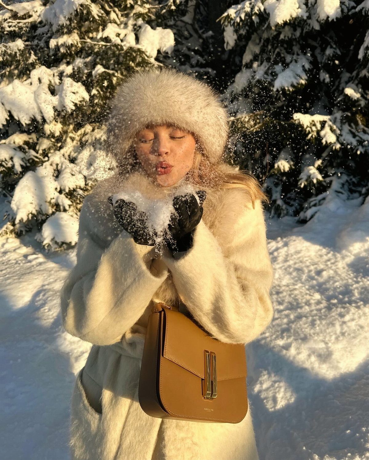 A woman in a white fuzzy coat and fur hat blowing snow in a winter forest.