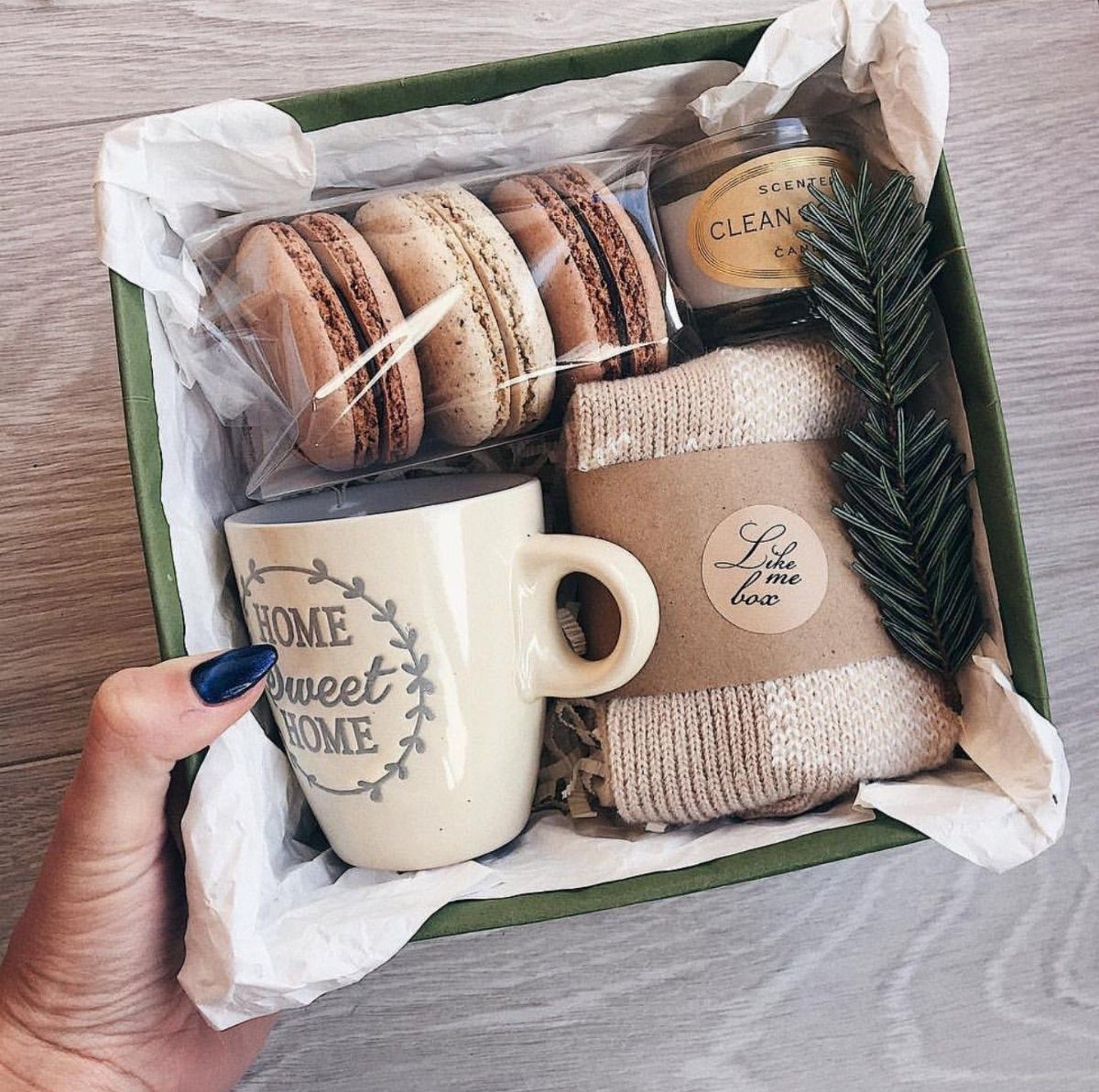 A hand holding a green gift box filled with macarons, a mug, and cozy textiles.