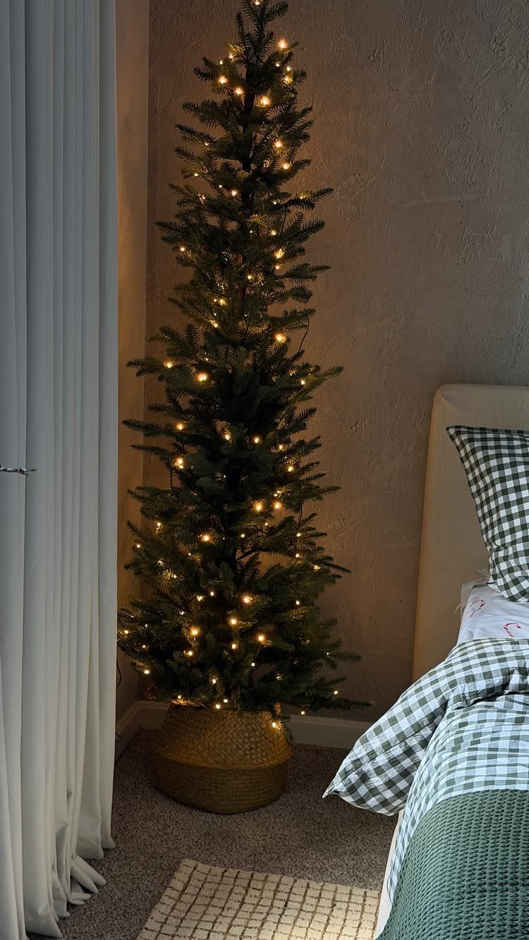A slim Christmas tree with warm lights standing in a gold basket in a bedroom.