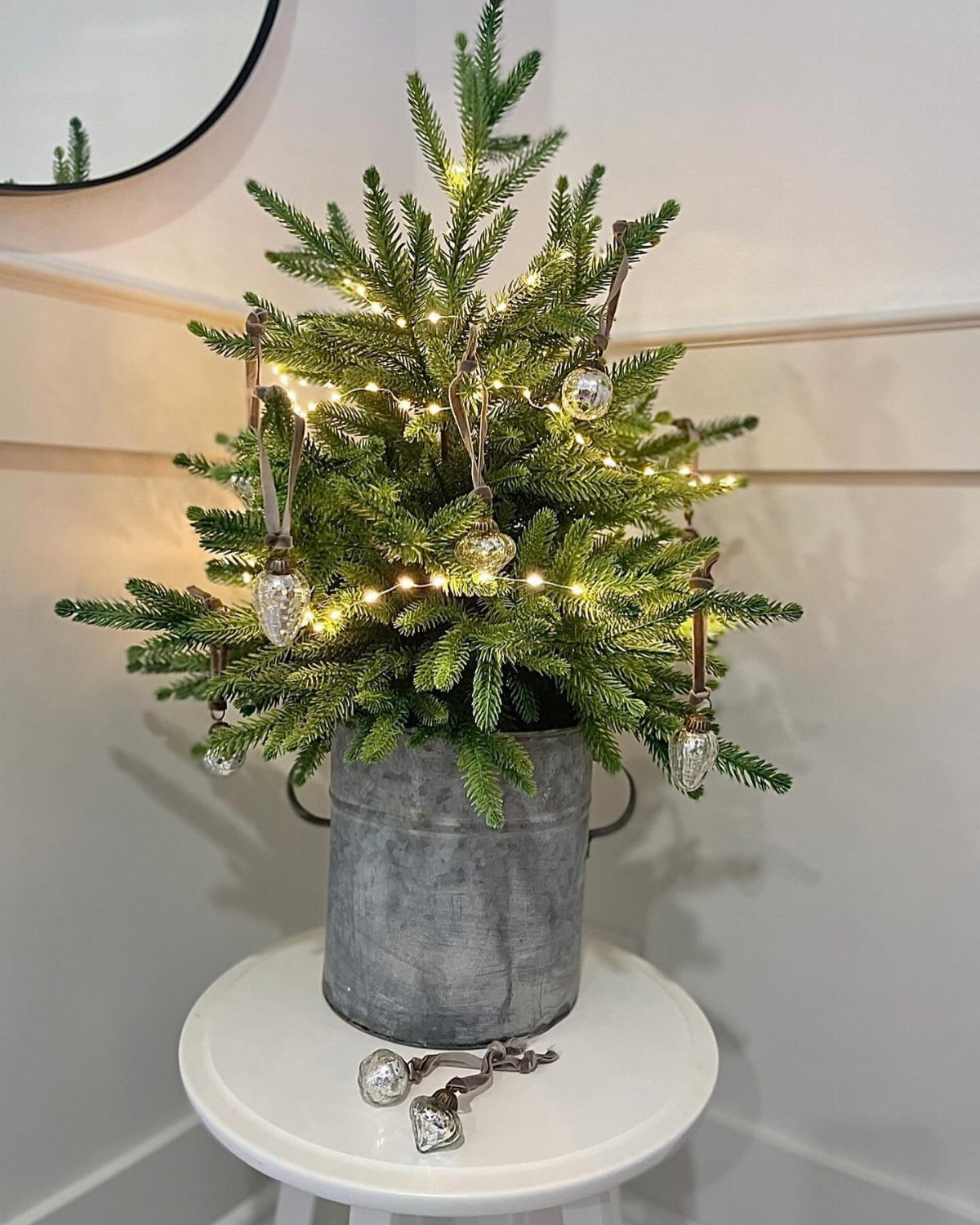 Small lit evergreen Christmas tree in a metal bucket on a white stool.
