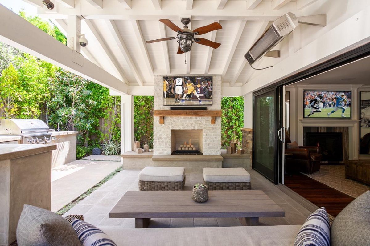 7 Back Porch Outdoor Kitchen Ideas for Covered Cooking 5 Covered patio with stone fireplace, television, ceiling fan, and lounge seating connecting to an indoor room.