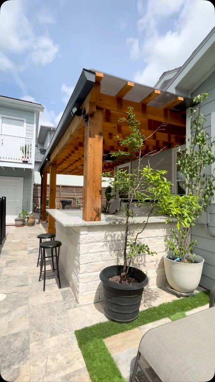 7 Back Porch Outdoor Kitchen Ideas for Covered Cooking 2 A stone outdoor kitchen island with bar stools situated under a large wooden pergola structure.