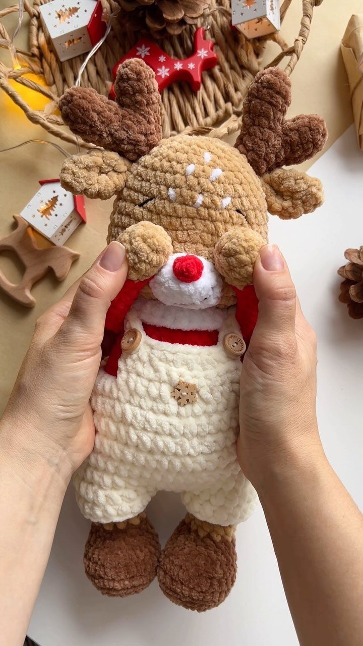 Hands holding a small, handmade crochet reindeer doll with brown antlers and a red nose.