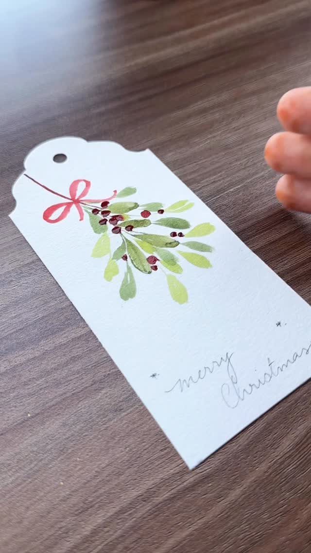 A white watercolor Christmas gift tag with mistletoe illustration on a wooden table.