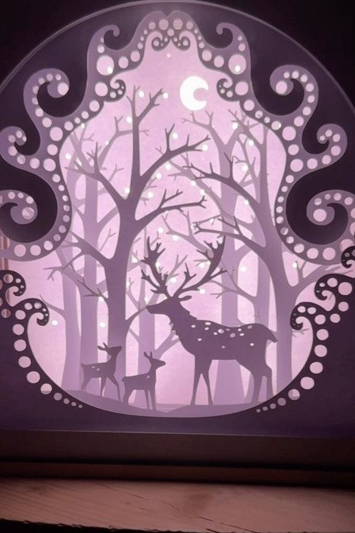 A glowing light shadow box depicting deer silhouettes in a forest under a crescent moon.