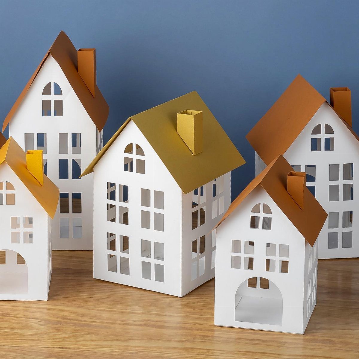 Small white paper houses with colorful roofs arranged on a wooden table against a blue background.