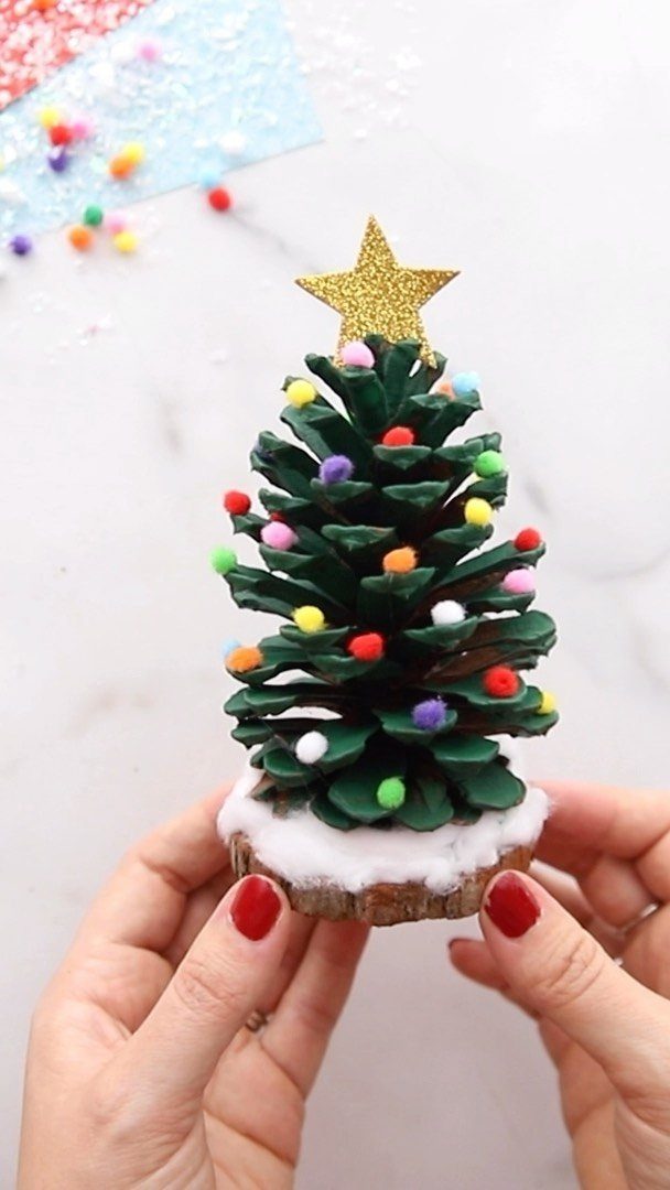Hands holding a pinecone decorated with colorful pom poms and a gold star topper.