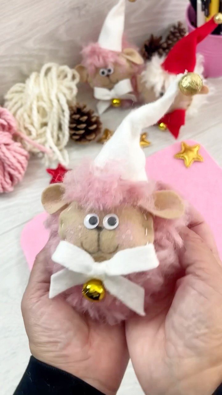 Hands holding a pink pom pom sheep ornament with a white hat and gold bell.