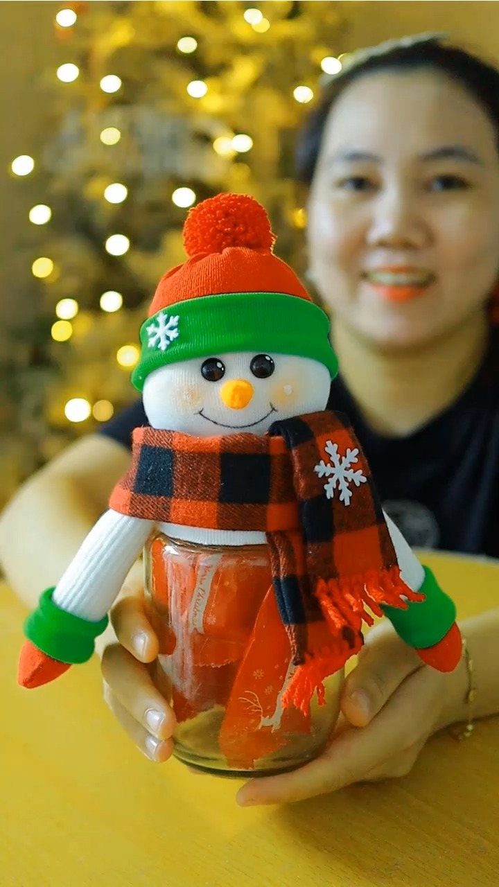 A person holding a snowman-themed mason jar filled with orange candies against a blurred Christmas tree.
