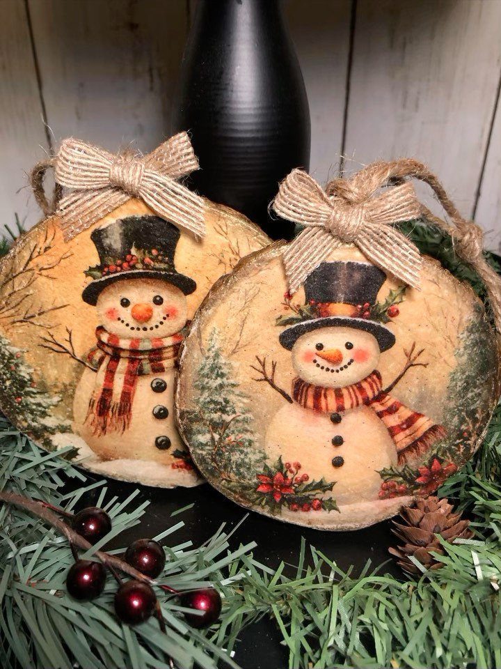 Two snowman painted wood slice ornaments with burlap bows resting in green pine branches.