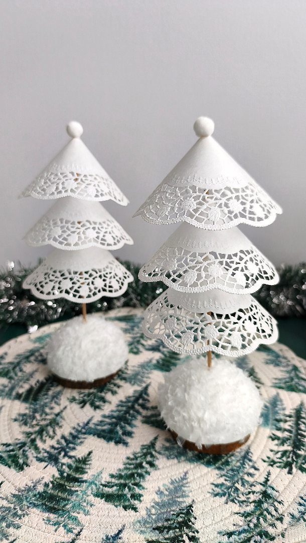 Two white tiered paper doily Christmas trees sitting on fluffy white bases.