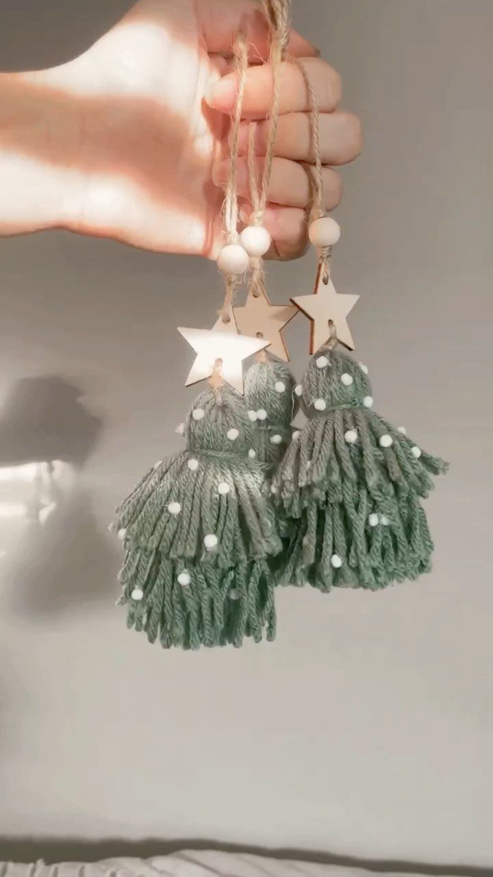 A hand holding three sage green yarn tassel ornaments with wooden stars and white beads.