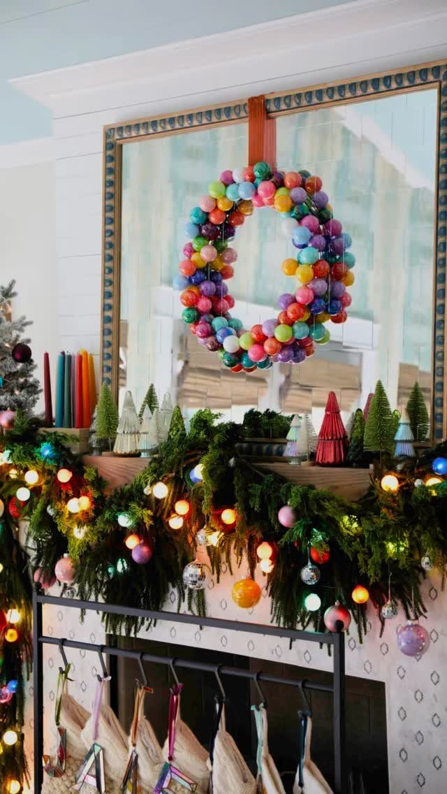 Christmas mantel decorated with green garland, colorful ornaments, and small decorative trees in front of a mirror.