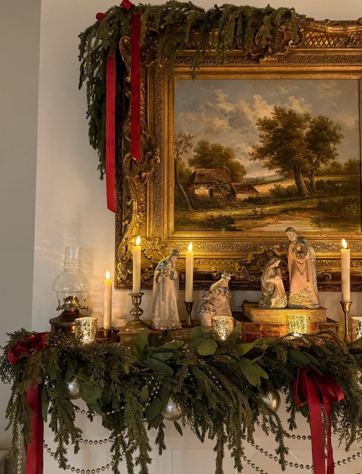Evergreen garland with red ribbons and angel figurines decorating a mantelpiece above a gold framed painting.