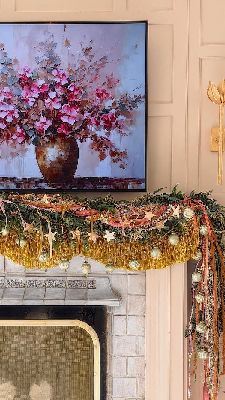 A decorated fireplace mantel featuring evergreen garland, gold tinsel, and pink ribbons under a floral painting.