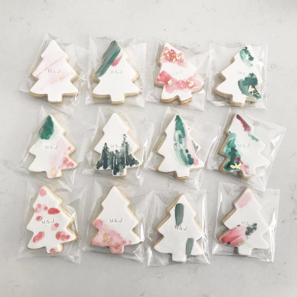 Twelve tree-shaped sugar cookies with watercolor painted icing designs, individually wrapped in clear plastic bags.