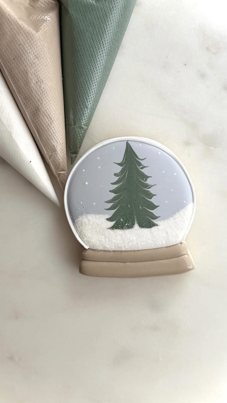 A snow globe decorated sugar cookie with a green tree and white snow on marble.