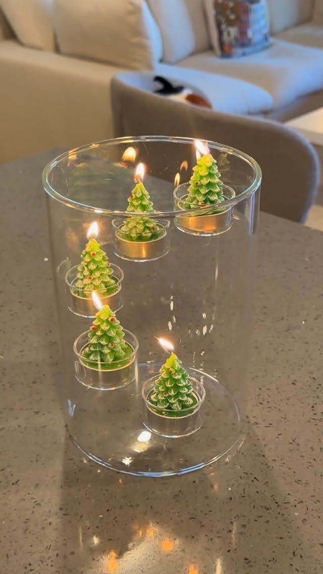 Small green tree-shaped candles floating in water inside a large glass cylinder vase.
