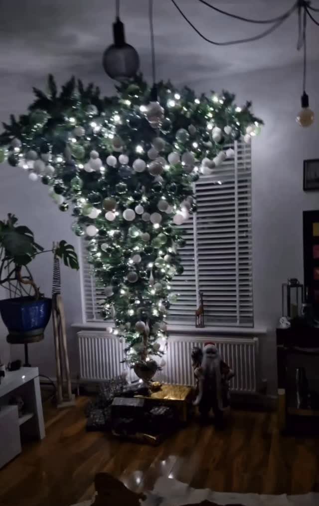 An upside-down Christmas tree decorated with white ornaments and lights hanging from a ceiling.