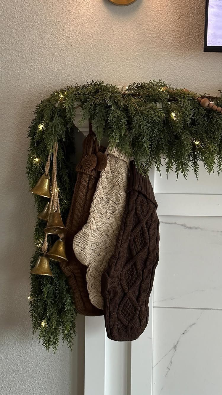 Knitted brown and cream Christmas stockings hanging from a lit evergreen garland on a white mantel.