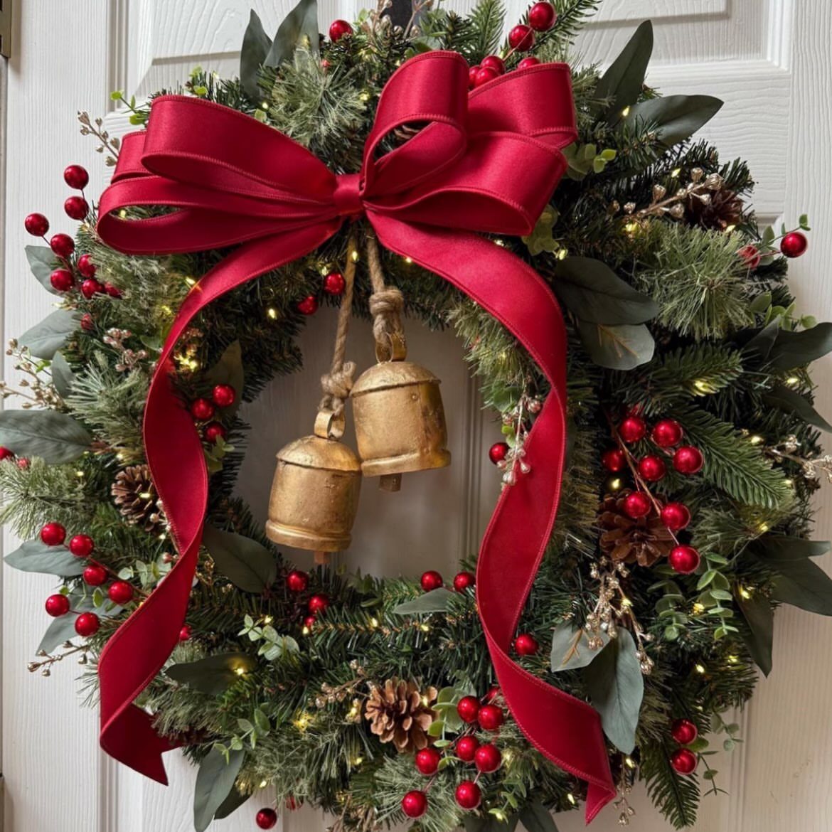 Green evergreen wreath decorated with red berries, pinecones, a large red velvet bow, and gold bells.