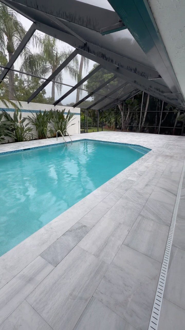 Light blue swimming pool surrounded by large grey rectangular tiles under a mesh screen enclosure.