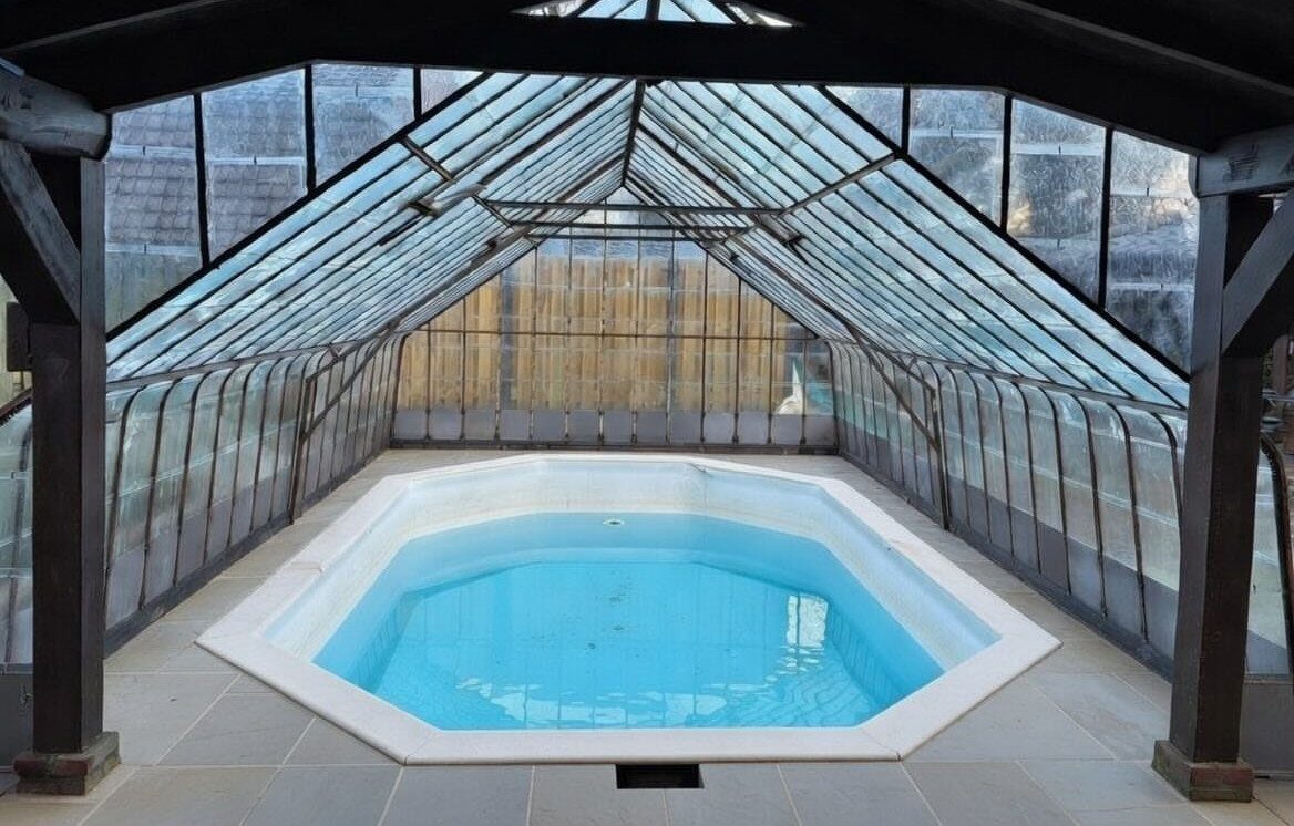 A small hexagonal pool enclosed by a structure with clear glass panels and a peaked roof.