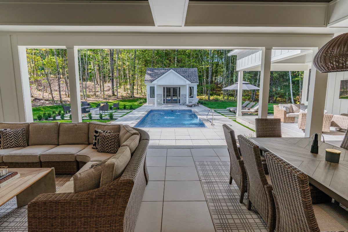 Covered patio with outdoor furniture overlooking a swimming pool and wooded backyard area.