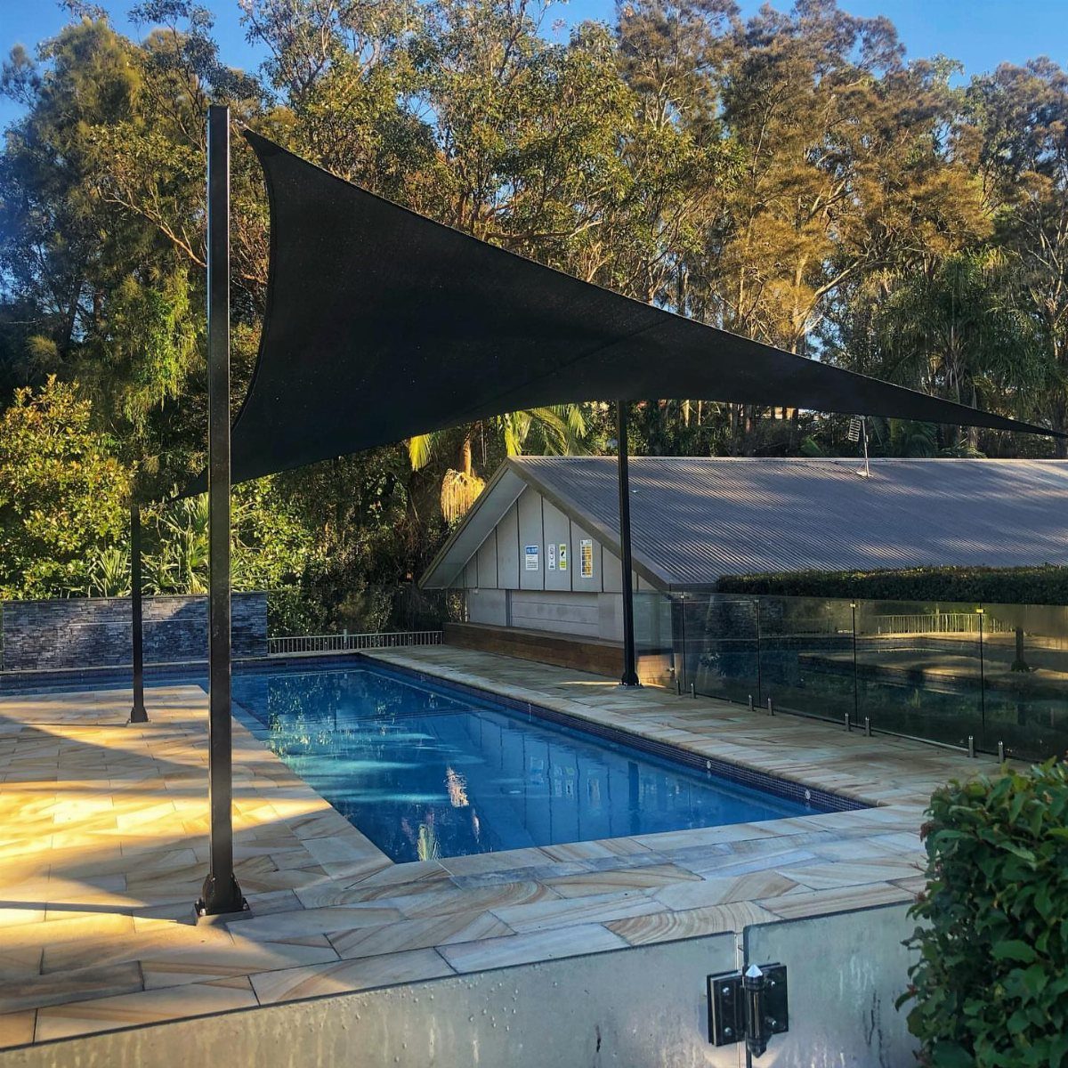 A black triangular shade sail stretched over a swimming pool with stone tiling and trees.