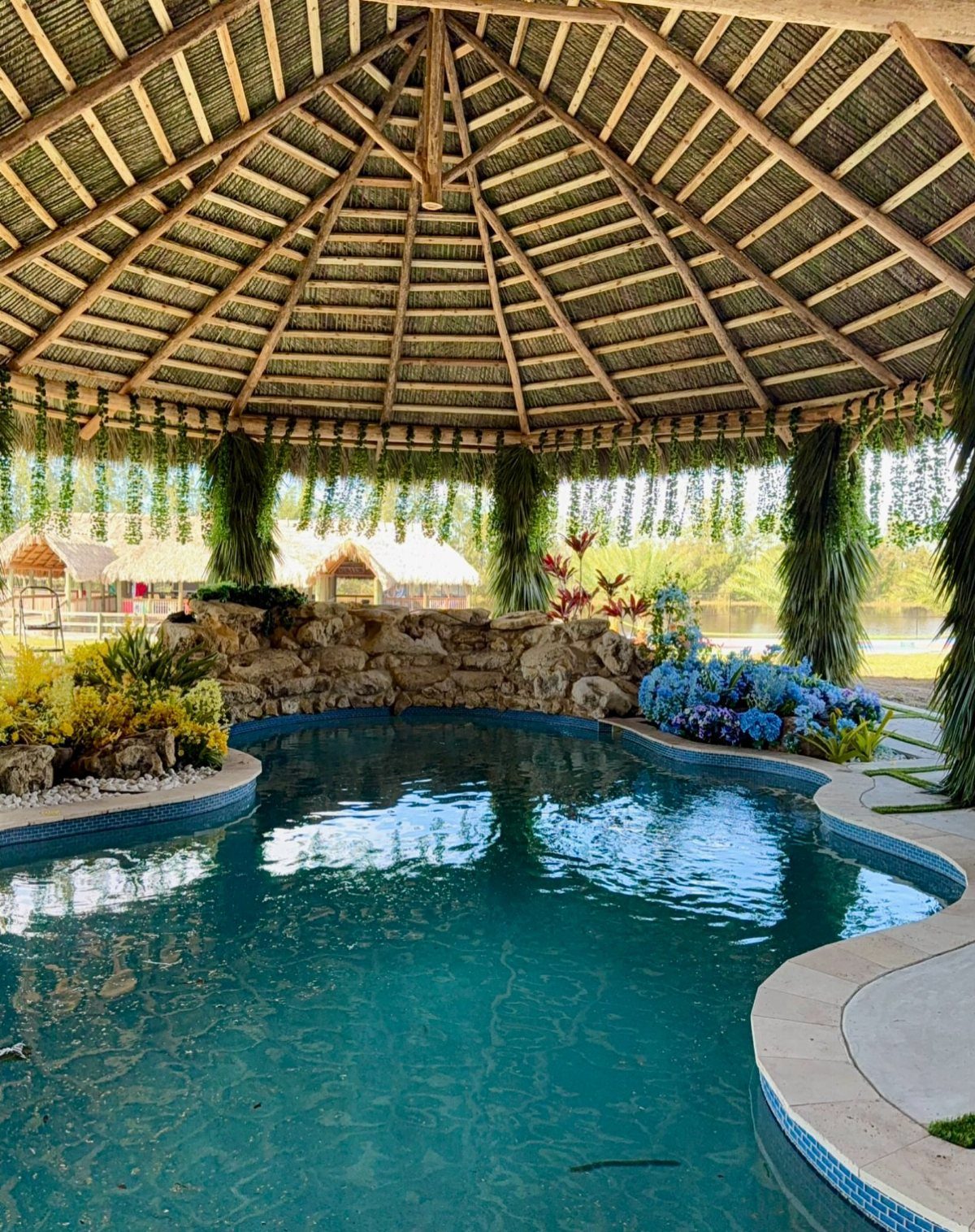 A large thatched palapa structure overlooking a bright blue swimming pool with lush tropical landscaping.