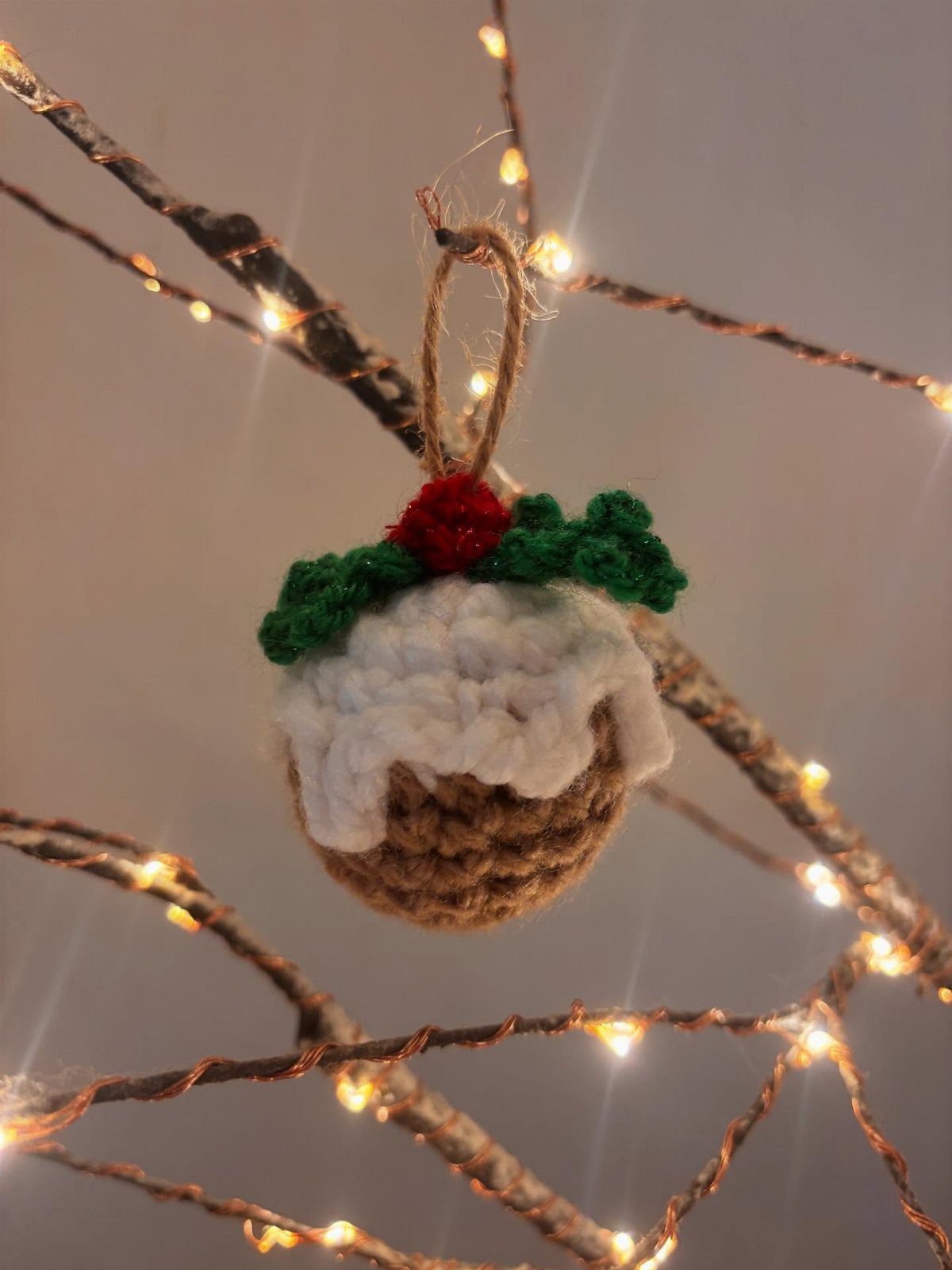 Crochet Christmas Ornaments: 19 Free Patterns for 2026 15 A crochet Christmas pudding ornament with white frosting and green holly leaves hanging on fairy lights.
