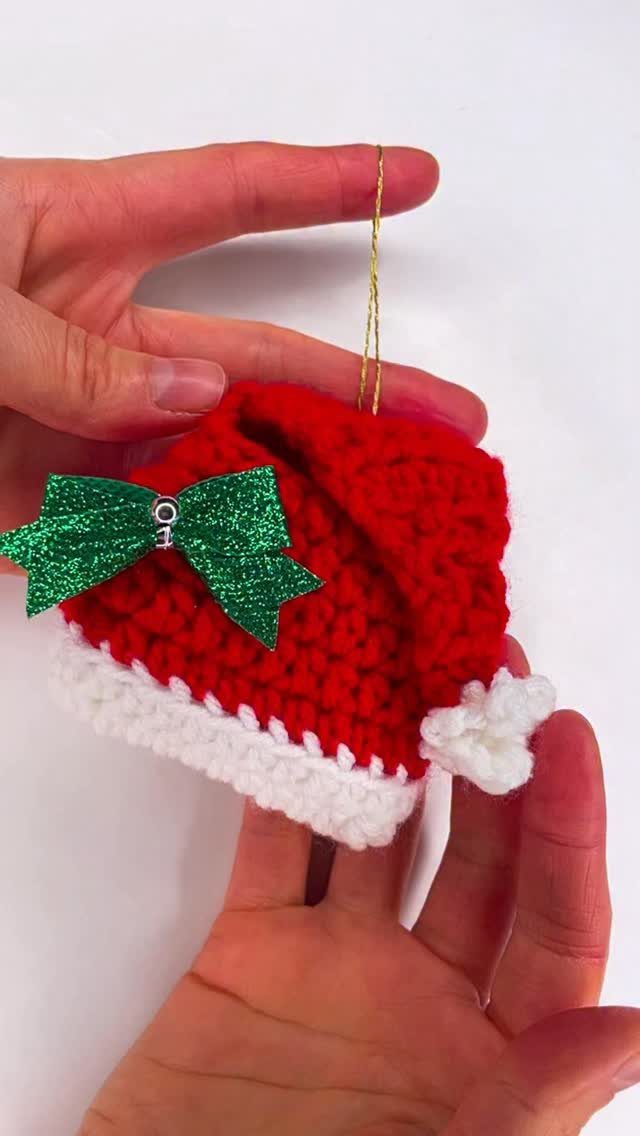Crochet Christmas Ornaments: 19 Free Patterns for 2026 7 Hands holding a small red and white crocheted Santa hat ornament with a green bow.