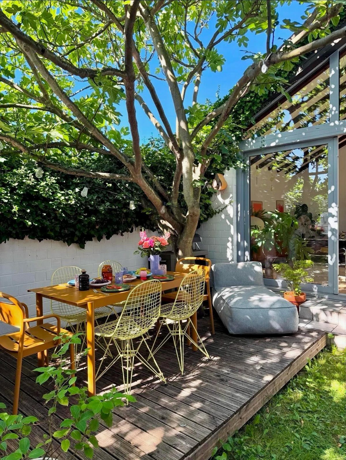 A yellow dining table with colorful dishes and wire chairs set on a wooden deck under a tree.