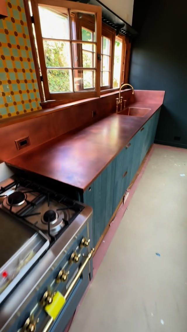 6 DIY Kitchen Countertops That Cost Less Than You Expect 5 Copper countertops installed on teal kitchen cabinets next to a window and a gas stove.