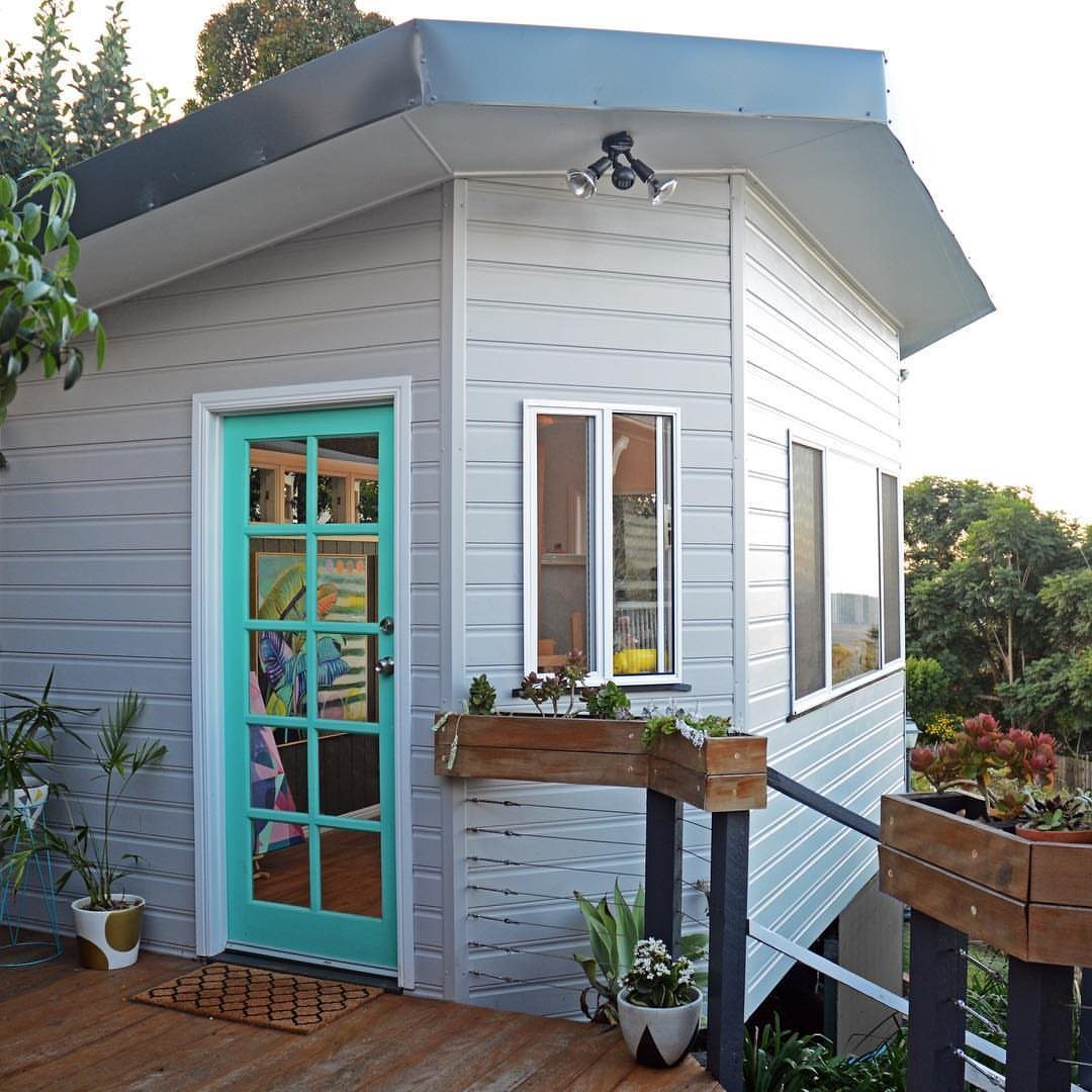 DIY Modern Shed: 19 Plans for Every Yard and Budget 4 A modern grey shed with a teal door and window frames on a wooden deck.