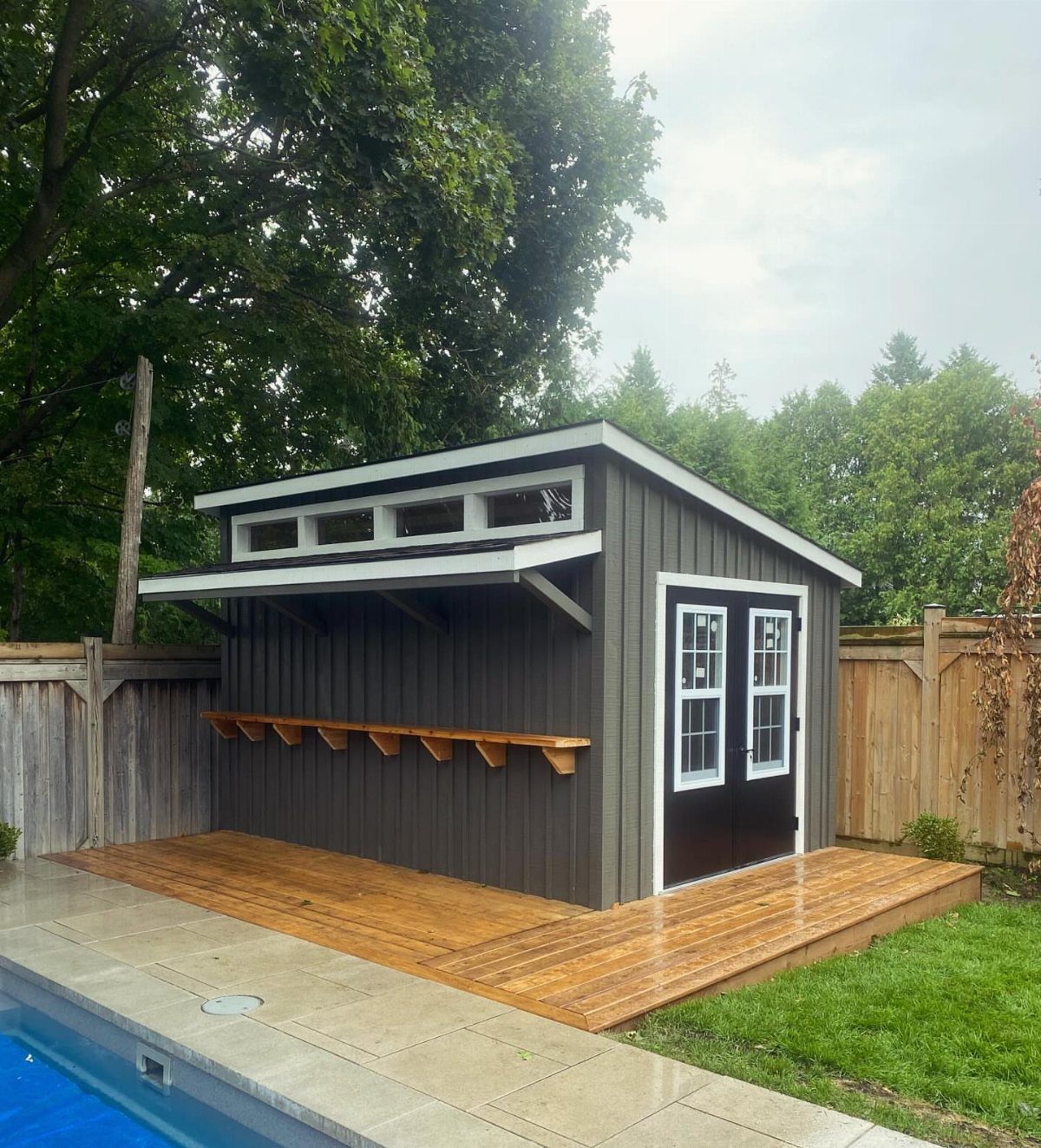DIY Modern Shed: 19 Plans for Every Yard and Budget 14 A dark grey modern wooden shed with white trim and a small wooden deck next to a pool.
