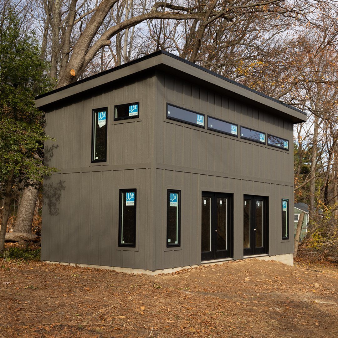 DIY Modern Shed: 19 Plans for Every Yard and Budget 12 A large two-story modern grey shed with black windows and doors in a wooded setting.