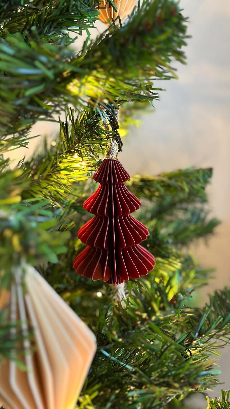 Red accordion-folded paper tree ornament hanging on a lit green Christmas tree branch.