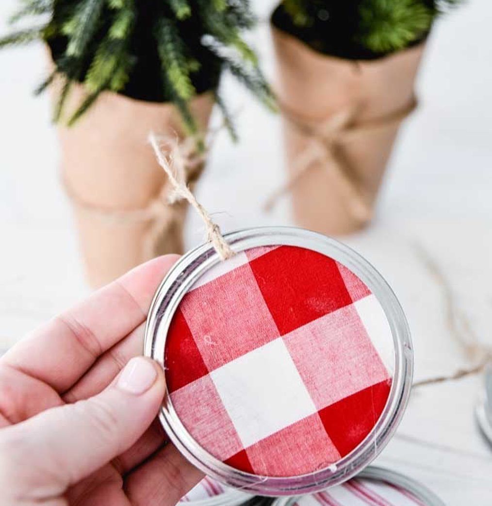 A hand holding a mason jar lid ornament featuring red and white checkered fabric and twine.