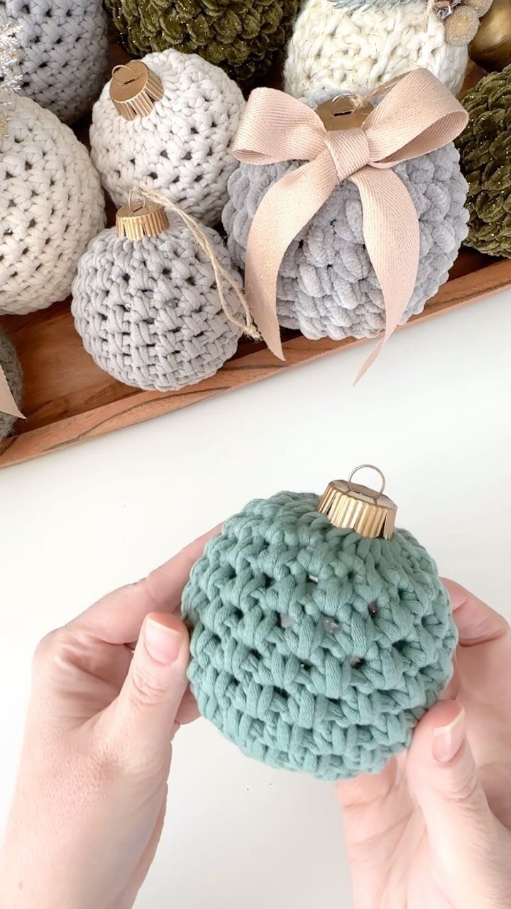 Hands holding a small teal crocheted ornament with several other colorful yarn ornaments in the background.