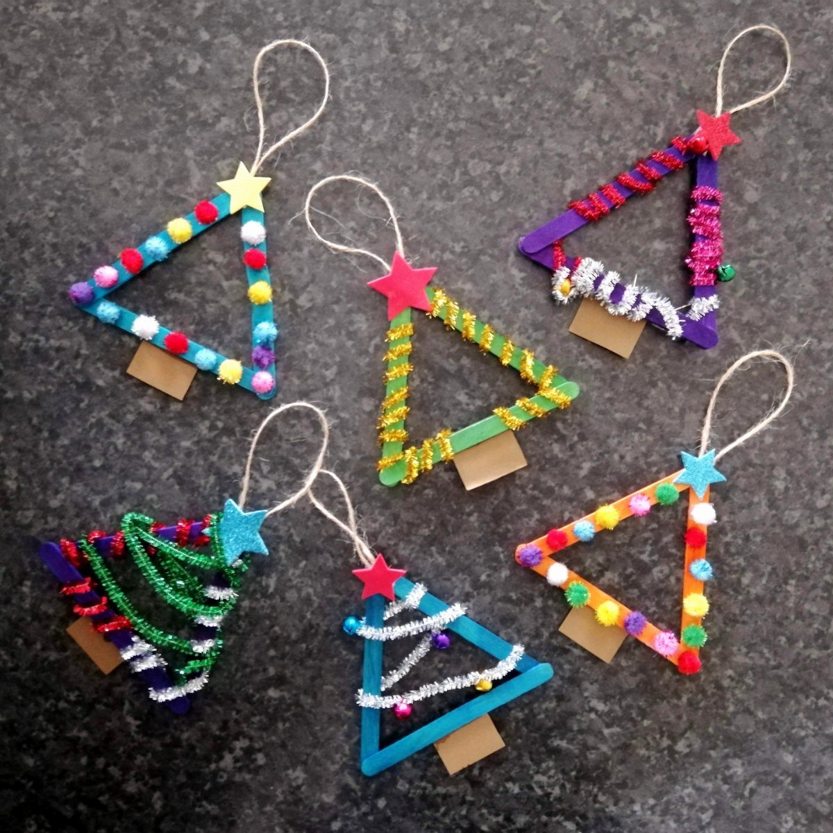 Six colorful popsicle stick Christmas tree ornaments decorated with glitter and pom-poms on a dark surface.
