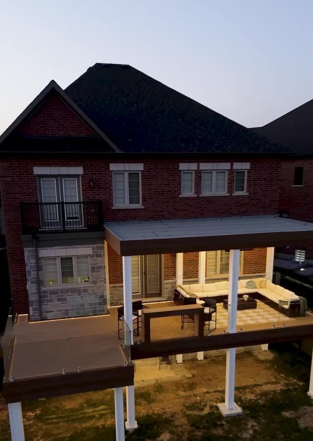 11 Elevated Deck Ideas: Multi-Level Looks, Storage & Rooftop Builds 1 A multi-level wooden deck with white pillars and glass railings attached to a brick house.