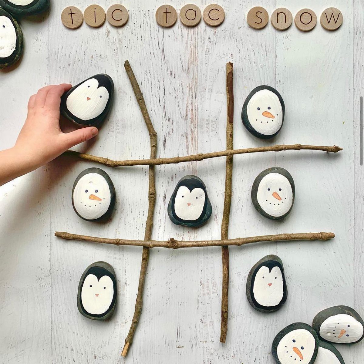 Fun Christmas Activities for Families: 18 Ideas With Photos 16 A hand placing a penguin-painted stone onto a twig tic-tac-toe board on white wood.