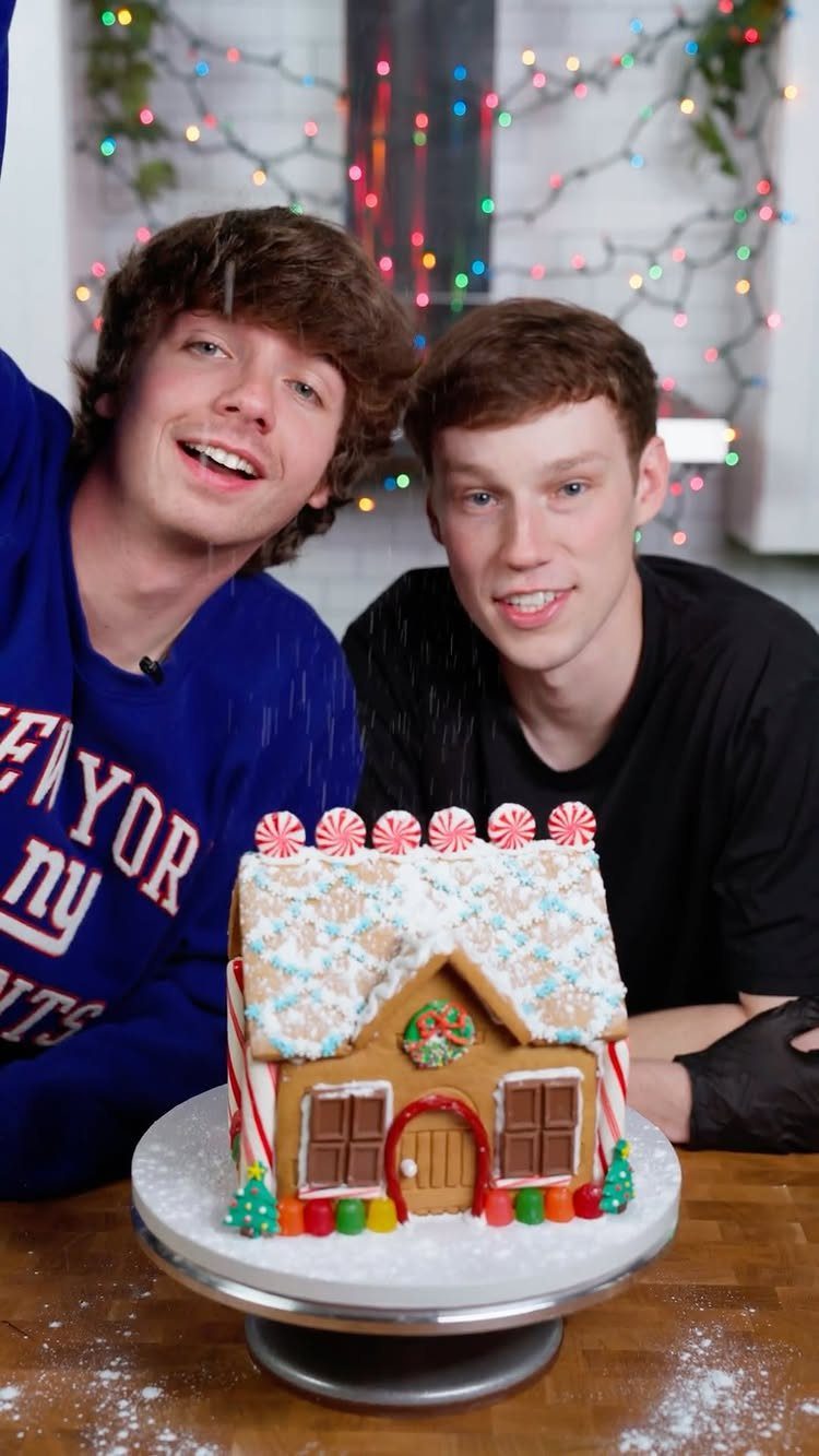 Fun Christmas Activities for Families: 18 Ideas With Photos 10 Two young men smiling behind a decorated gingerbread house with colorful candy decorations.