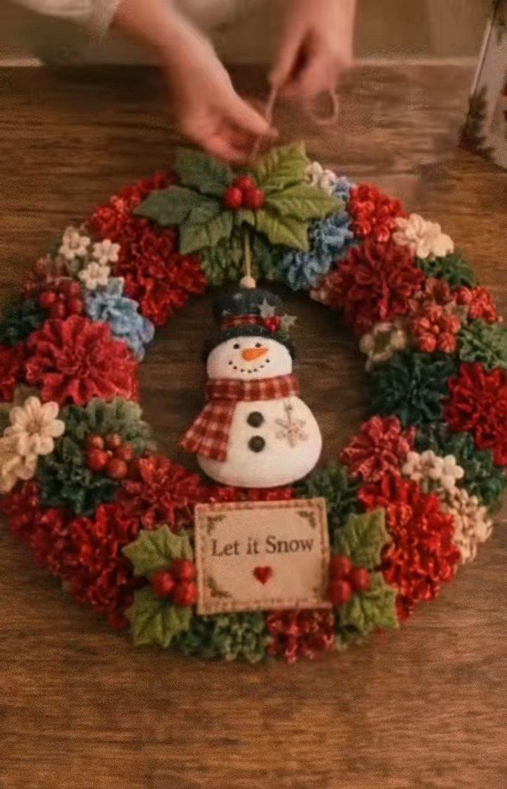 A colorful fabric flower wreath featuring a small snowman ornament and a Let it Snow sign.