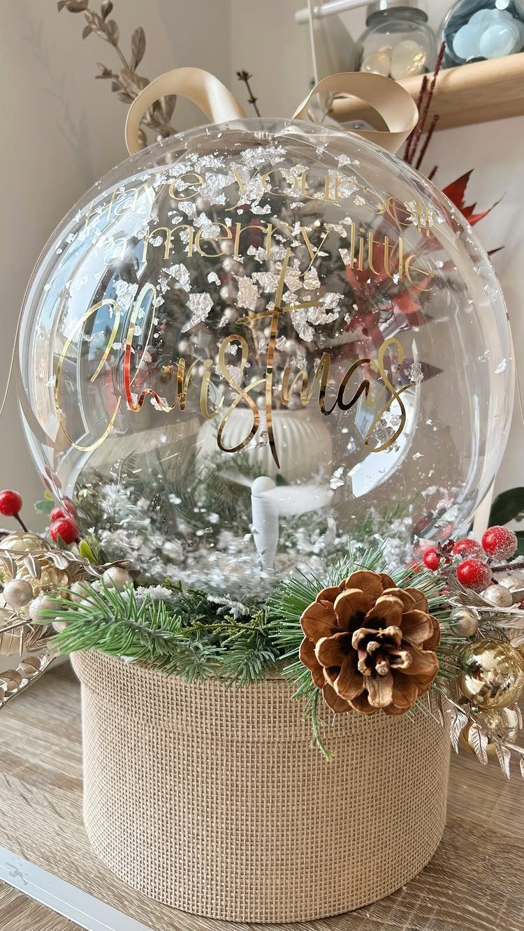 A clear spherical snow globe decorated with pine branches, red berries, and a pinecone on burlap.
