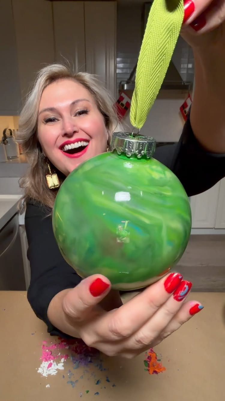 Smiling woman holding a large green marbled Christmas ornament with colorful crayon shavings below.